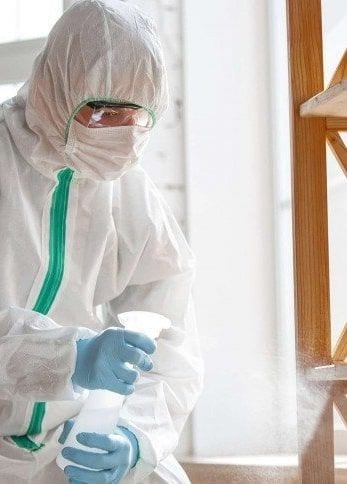 A technician in a white protective suit, goggles and mask spraying beside a window